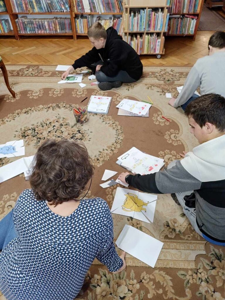 Na dywanie siedzi trzech uczestników zajęć i bibliotekarka. Przed nim karty pracy -  złożone z pociętych kartek kolorowe potworki. W tle regały z książkami.