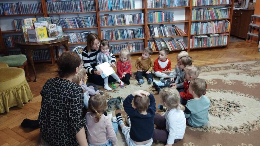 Grupa dzieci, opiekunka i bibliotekarka siedzą, w kole, na dywanie. 