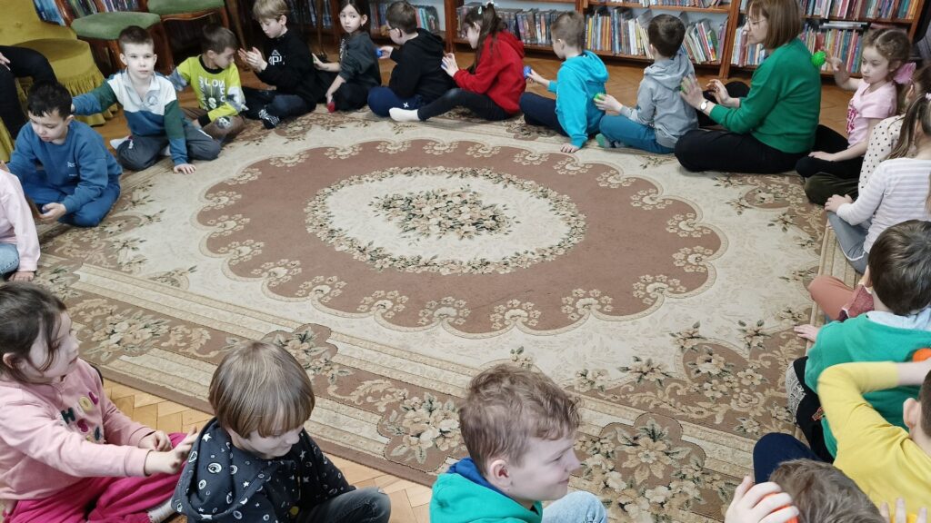 Dzieci siedzą na dywanie, tworząc koło. Każde z nich masuje piłeczką plecy sąsiada.