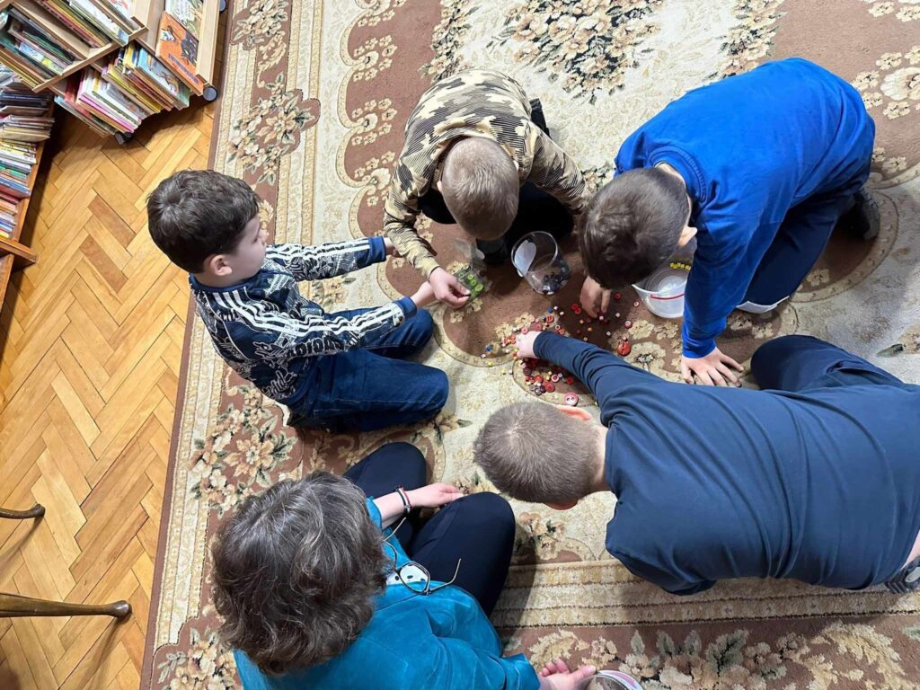4 dzieci i bibliotekarka siedzi na dywanie. Przed dziećmi stoją 4 plastikowe pudełka oraz leżą na dywanie kolorowe guziki. Dzieci wkładają guziki wg. kolorów do  pudełek.