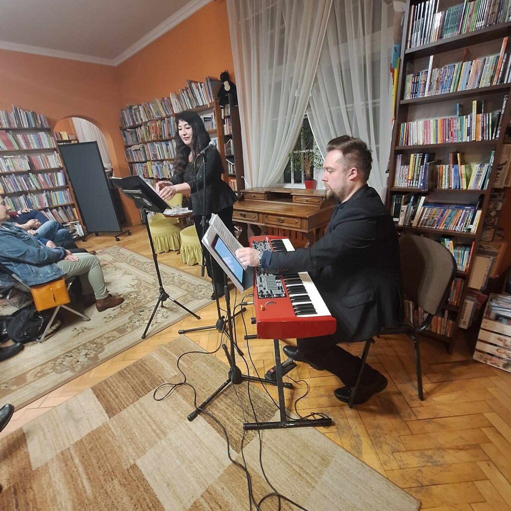 Andrzeja Mikulski siedzi na krześle i przekłada nuty. Przed mężczyzną keyboard. Obok mężczyzny Stoi Ewa Mikulska. 