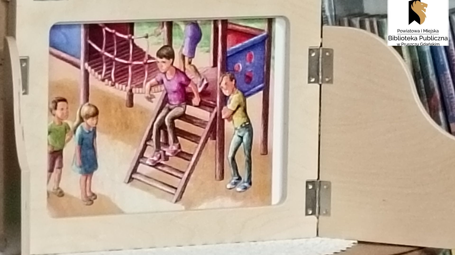 Fragment otwartej skrzynki kamishibai, z kolorową planszą przedstawiającą dzieci bawiące się na placu zabaw.