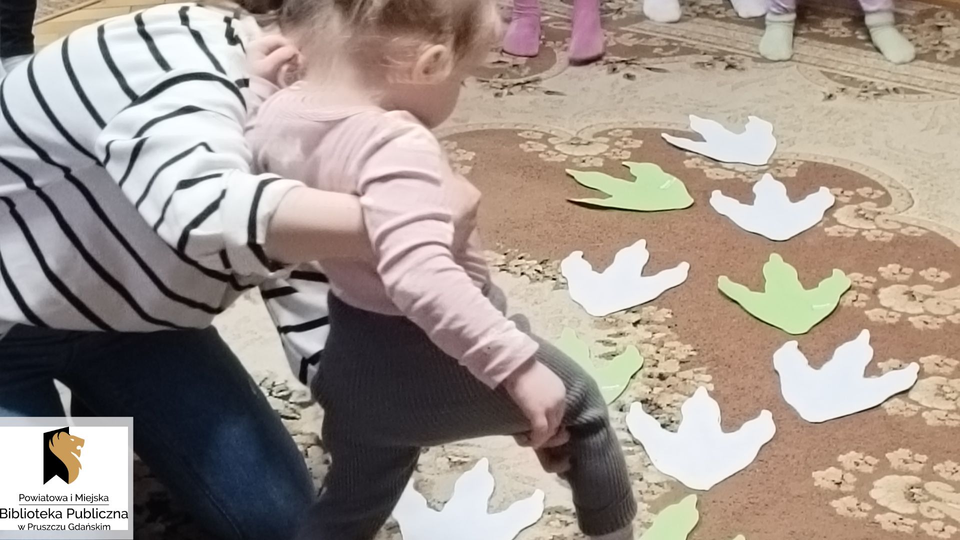 Mała dziewczynka, podtrzymywana przez mamę, idzie po śladach dinozaura, wyciętych z papieru