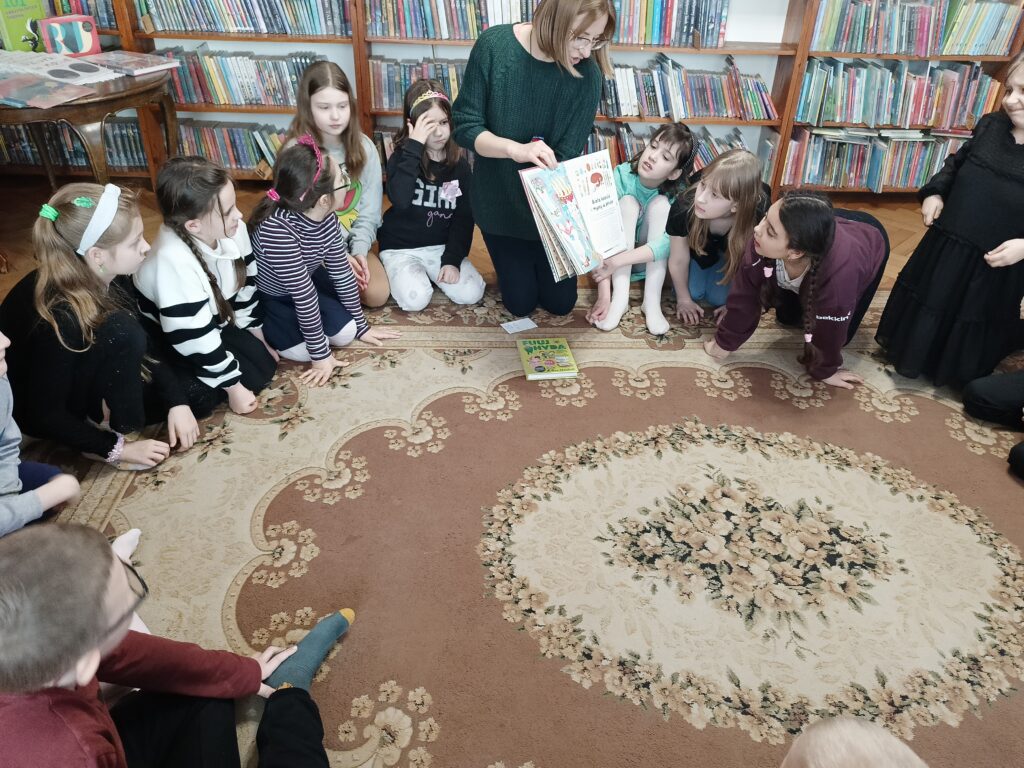 Na dywanie w okręgu siedzą dzieci. Miedzy nimi bibliotekarka, która pokazuje ilustrację z książki.