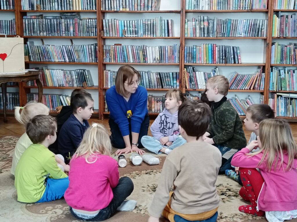 Kilkoro dzieci i bibliotekarka siedzą na dywanie. Przed kobietą leżą trzy rodzaje soli w opakowaniach.