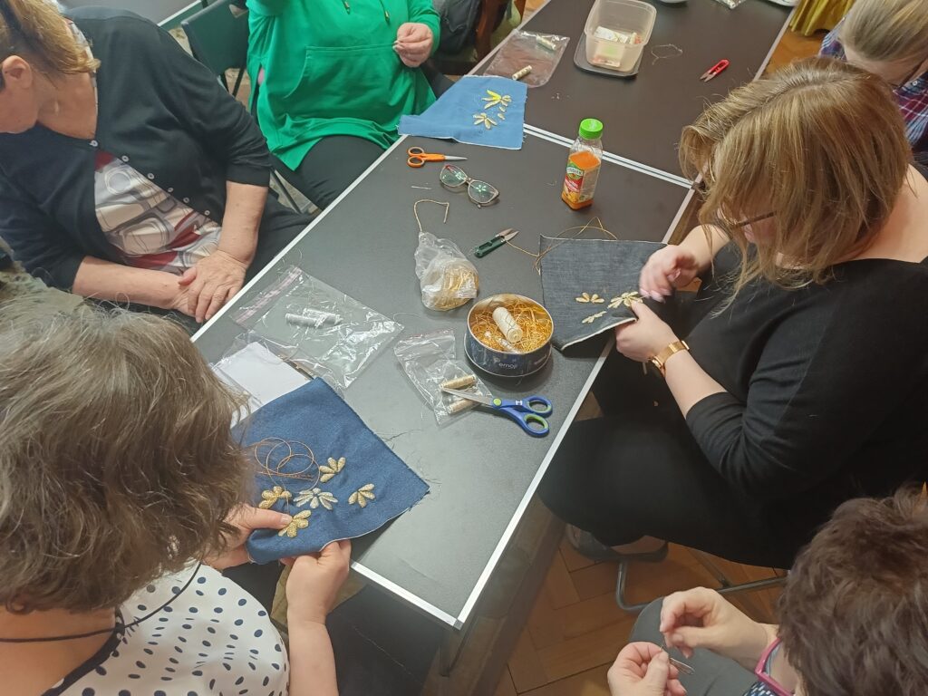 Kobiety siedzą przy stołach, haftują złotymi i srebrnymi niciami płatki kwiatów na kawałkach materiału jeansowego