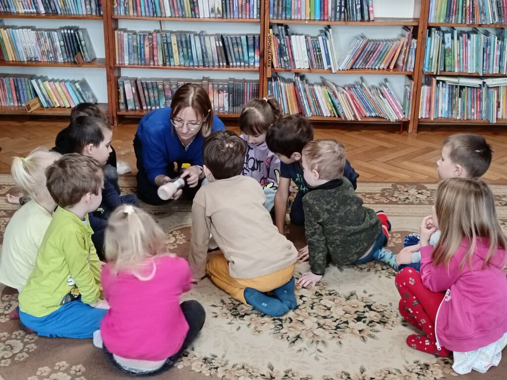 Kilkoro dzieci siedzi na dywanie. Bibliotekarka wskazuje na sól w młynku, który trzyma w rękach.
