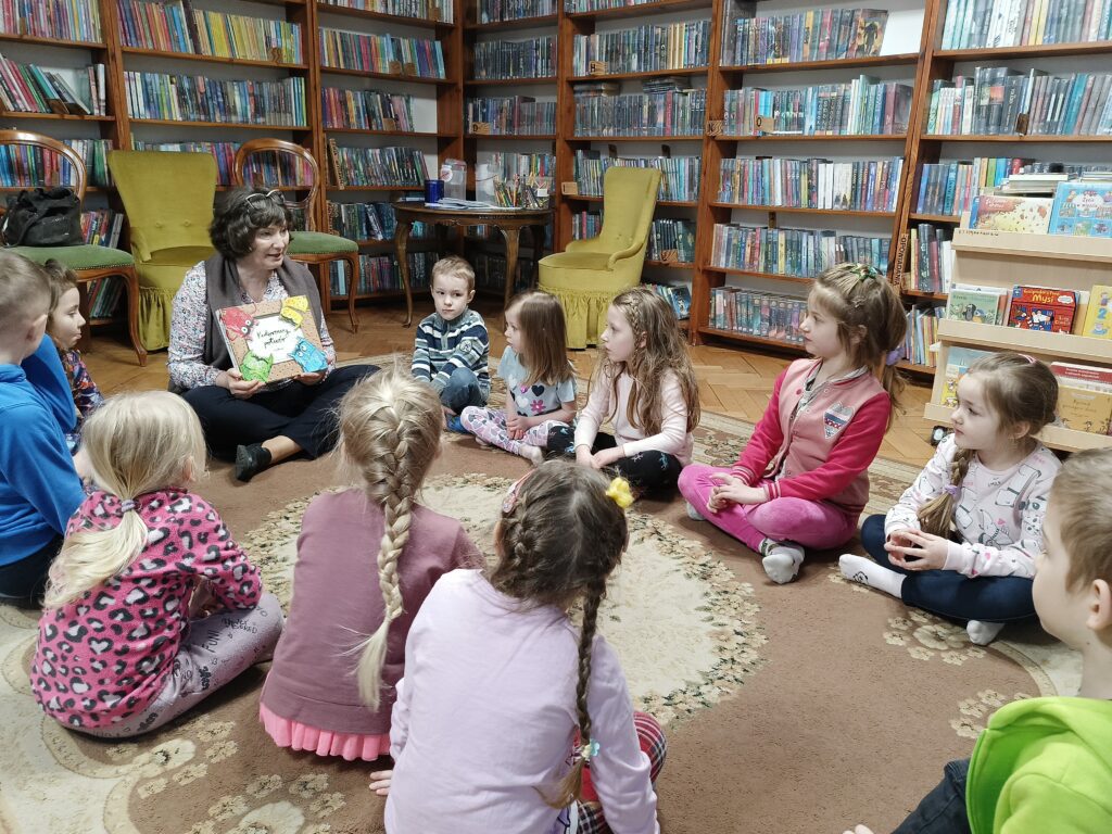 Kilkoro dzieci i bibliotekarka siedzą na dywanie. Kobieta trzyma książkę Kolorowy potwór