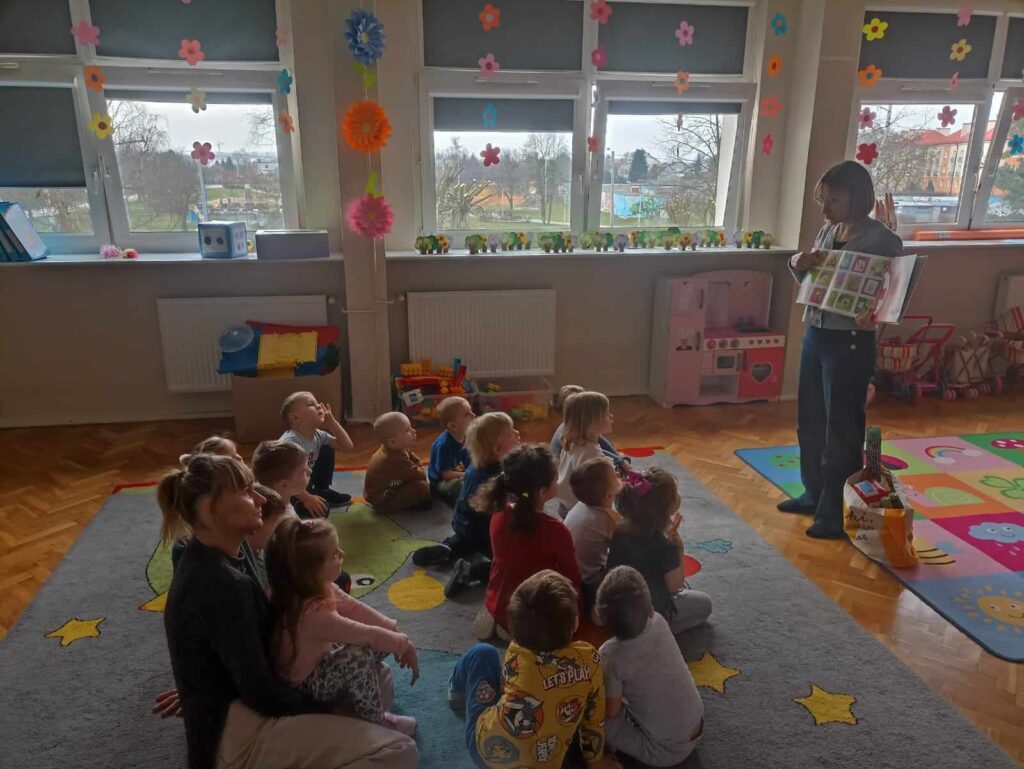 Bibliotekarka stoi przed siedzącymi na dywanie dziećmi. W ręku trzyma otwarta książkę.
