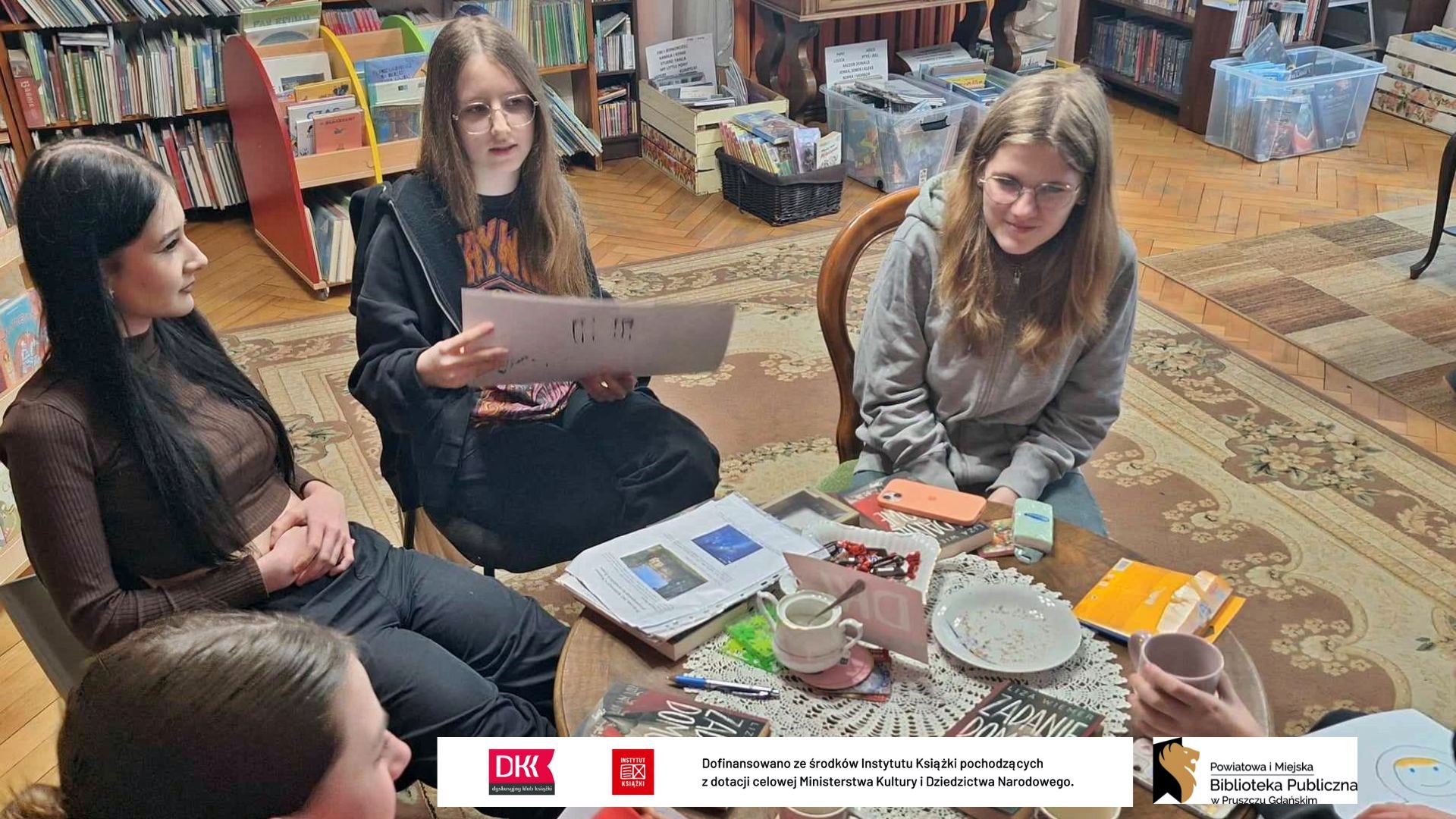 Przy stoliku siedzą 3 dziewczyny z Dyskusyjnego Klubu Książki dla młodzieży. Jedna z dziewczynek trzyma w rękach dużą kartkę i czyta. Na stoliku książki pt. Zadanie domowe.