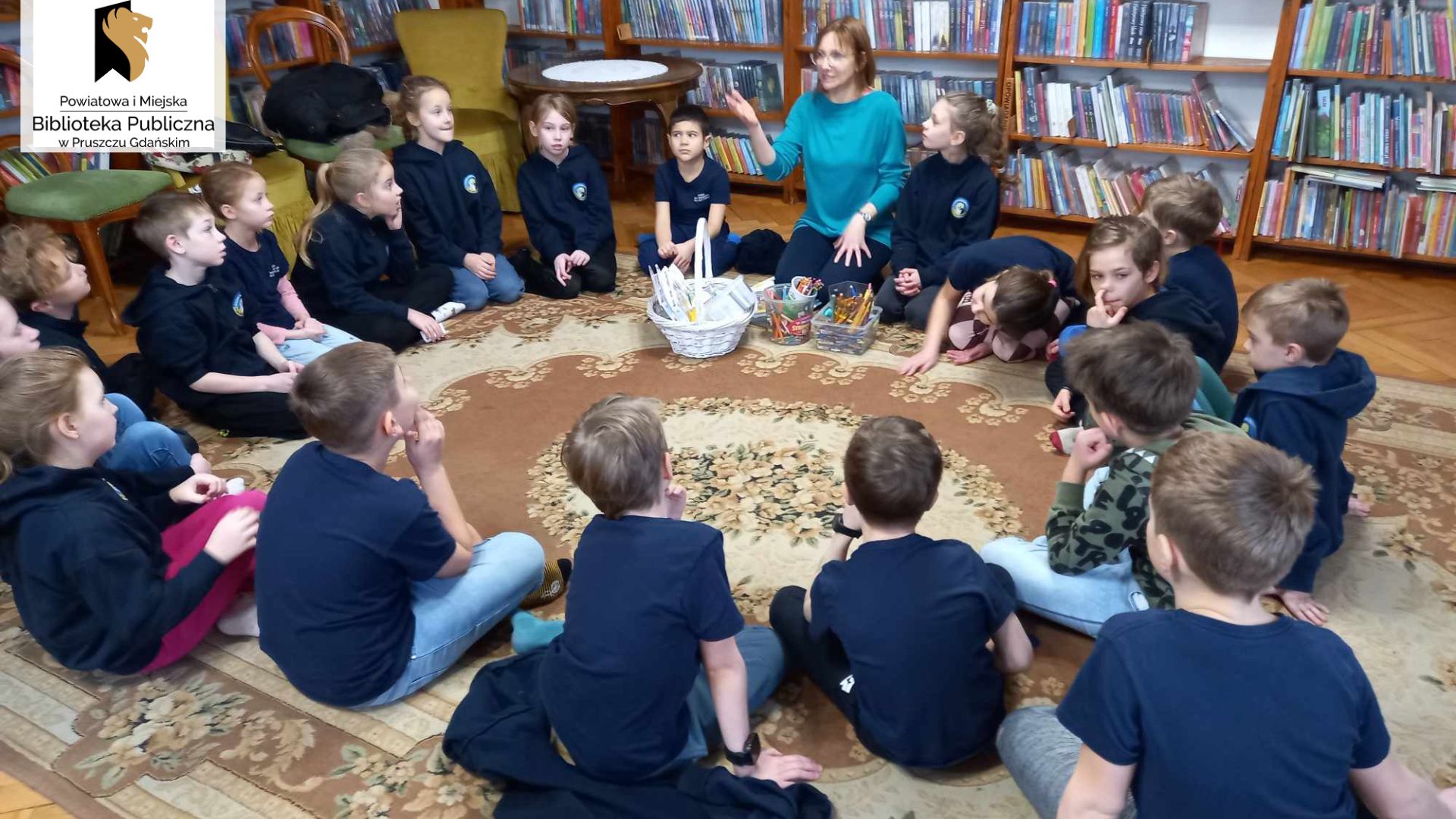 Na dywanie, w kole,, siedzą uczniowie i patrzą na siedząca z nimi bibliotekarkę. Przed nią koszyk z materiałami piśmienniczymi.