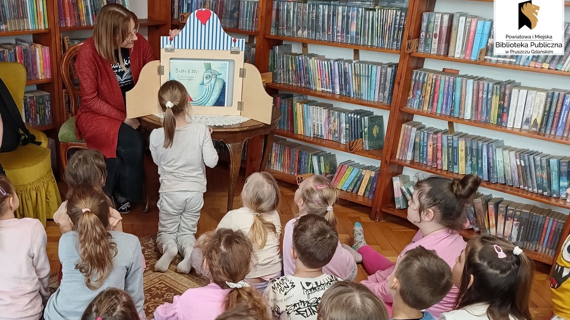 Dzieci siedzące na dywanie patrzą w stronę bibliotekarki prezentującej teatrzyk kamishibai.