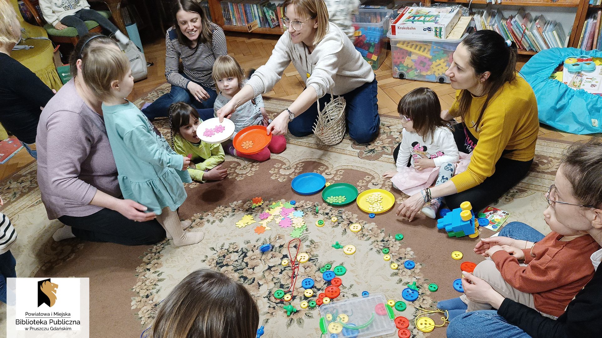 Na dywanie siedzą dzieci i rodzice. Lężą kolorowe talerzyki, rozsypane kolorowe guziki. Bibliotekarka wyciąga ręce z dwoma talerzykami do stojącego dziecka.