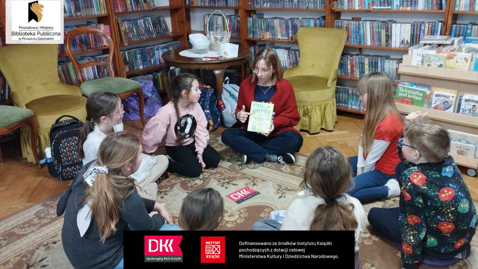 Dzieci i bibliotekarka siedzą na dywanie, w koło. Kobieta prezentuje książkę.