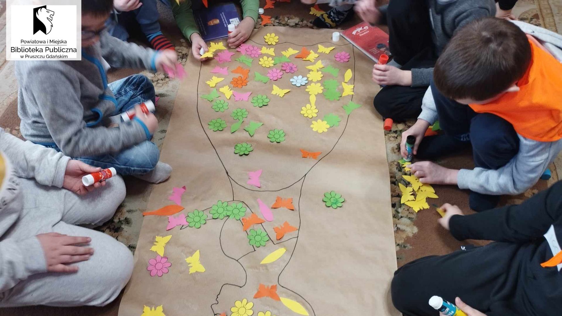 Grupa dzieci na dywanie wykleja motylkami i kwiatkami namalowaną na szarym papierze postać kobiety