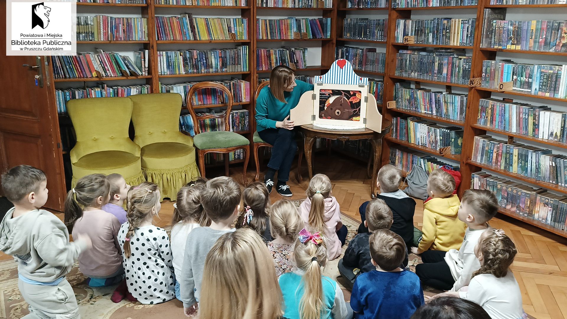 Dzieci siedzą tyłem na dywanie. Przed nimi, na krześle siedzi bibliotekarka. Obok niej otwarta skrzynka teatrzyku kamishibai