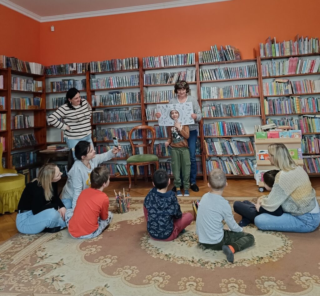 Uczestnicy spotkania siedzą na dywanie. Bibliotekarka trzyma otwartą książkę, z dziurą w środku, przez która zagląda chłopiec.