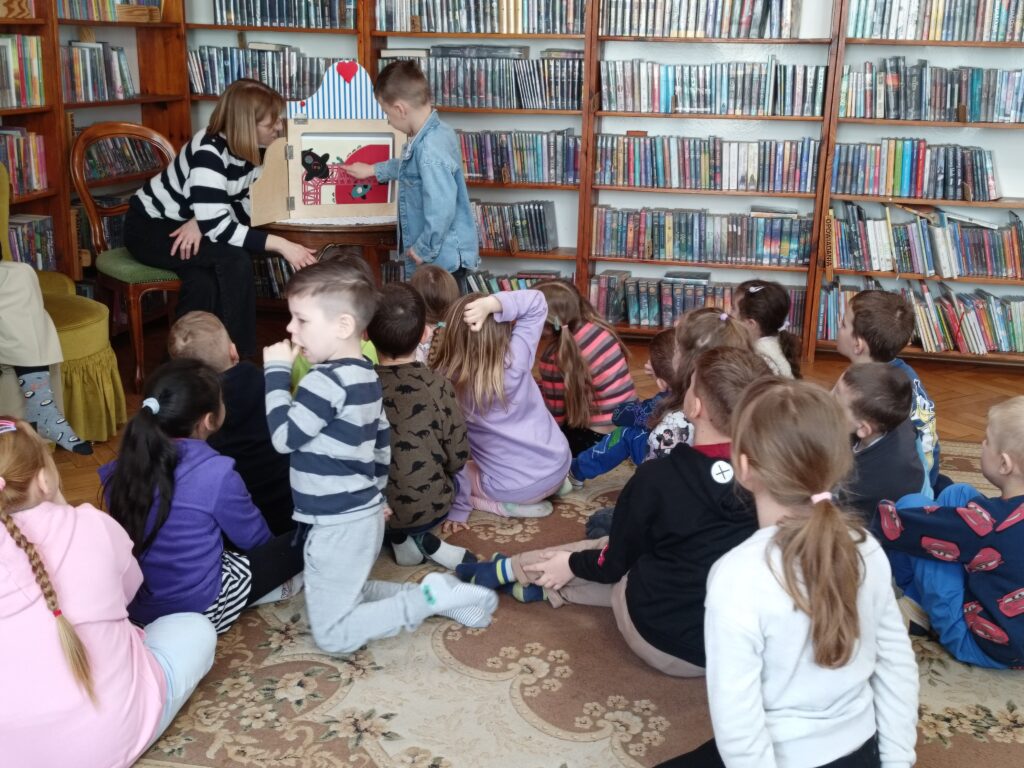 Dzieci siedzące na podłodze. Bibliotekarka i chłopiec przyglądają się ilustracji teatrzyku kamishibai.