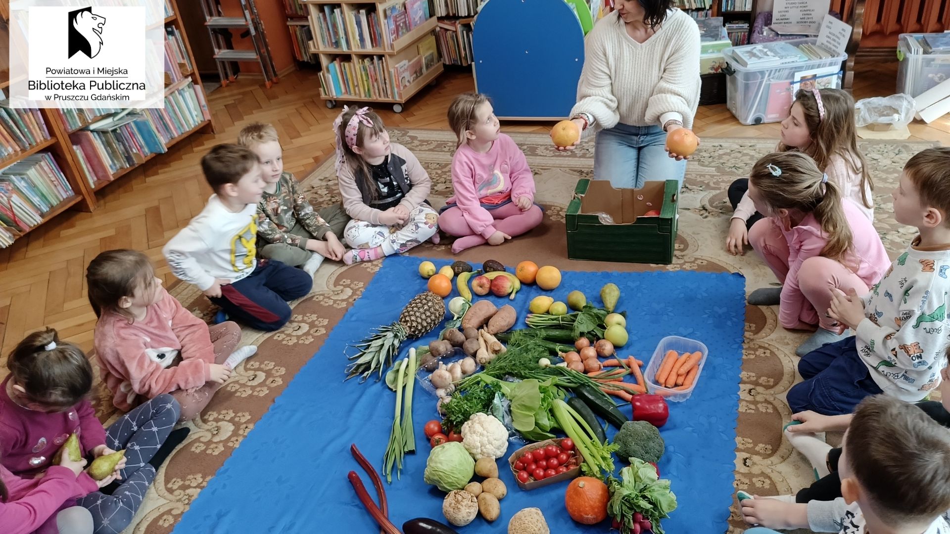 Dzieci siedzące na dywanie i Renata Szordykowska oglądją wspólnie warzywa i owoce.