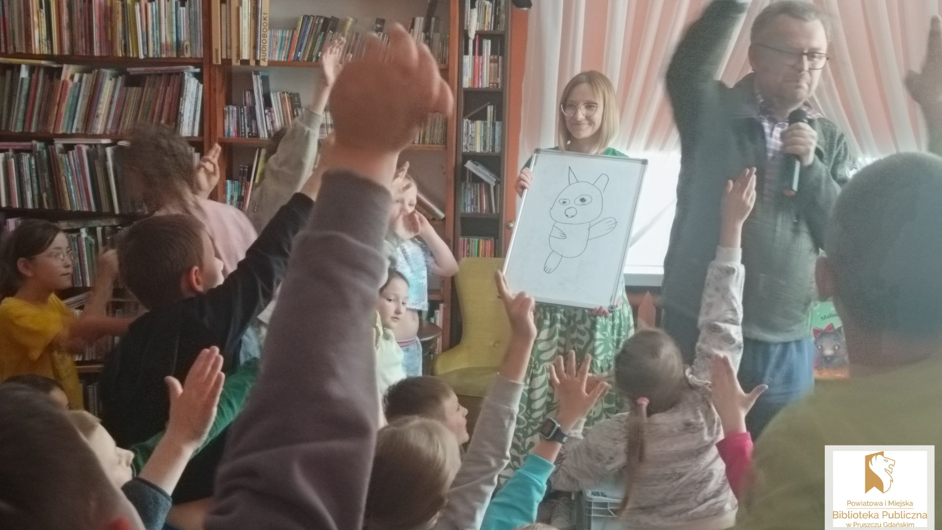 Marcin Kozioł i kobieta stoją przed dziećmi unoszącymi do góry ręce.