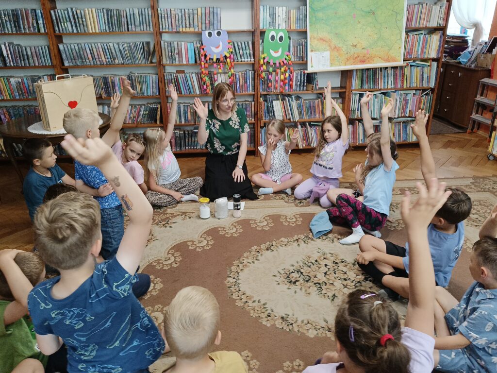 Grupa dzieci i bibliotekarka siedzą na dywanie.  Przed kobietą leżą pojemniki z solą.