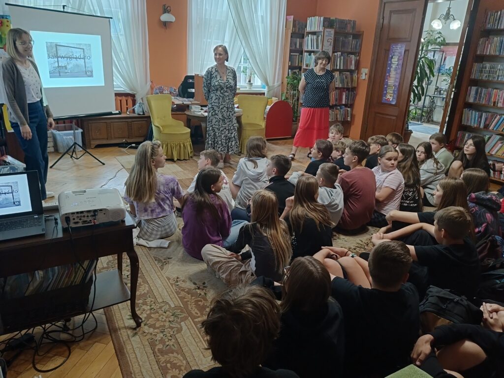 Autorka i bibliotekarki przed zgromadzoną młodzieżą, siedzącą na podłodze.