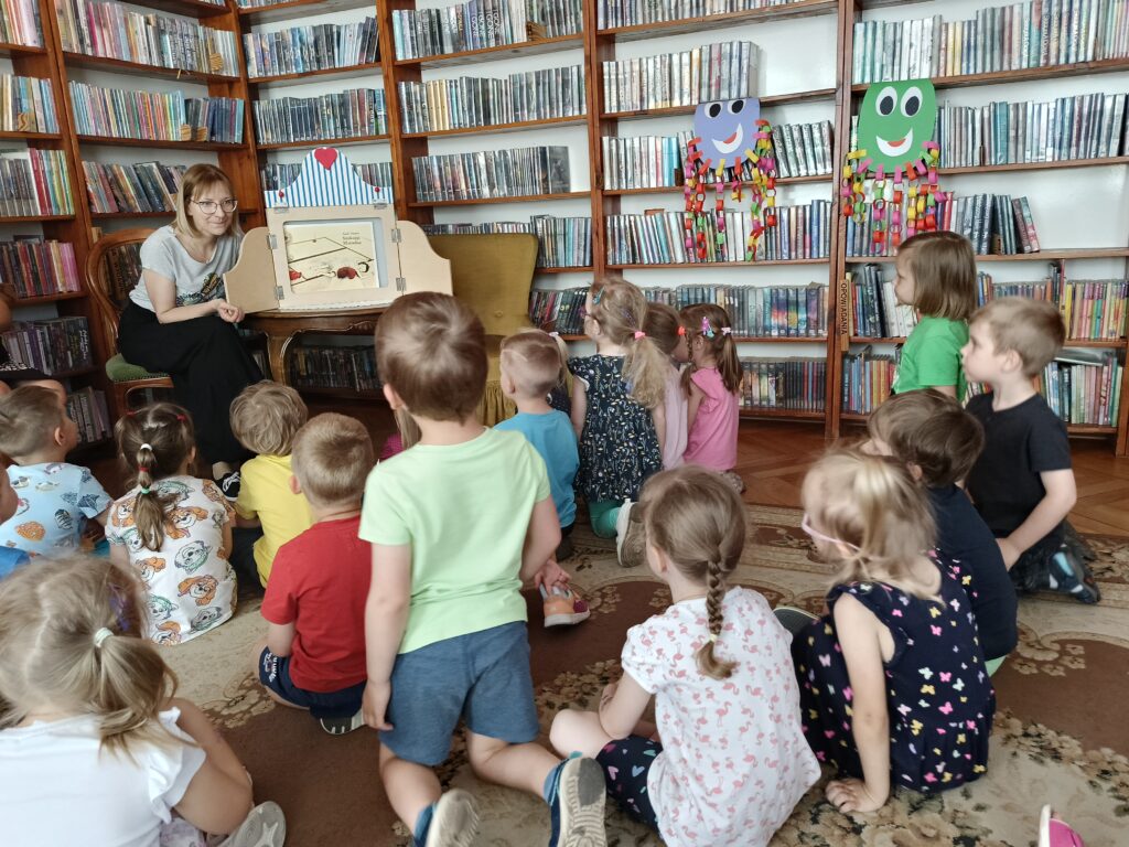 Bibliotekarka siedząca przy otwartej skrzynce do teatrzyku papierowego i dzieci siedzące na podłodze.