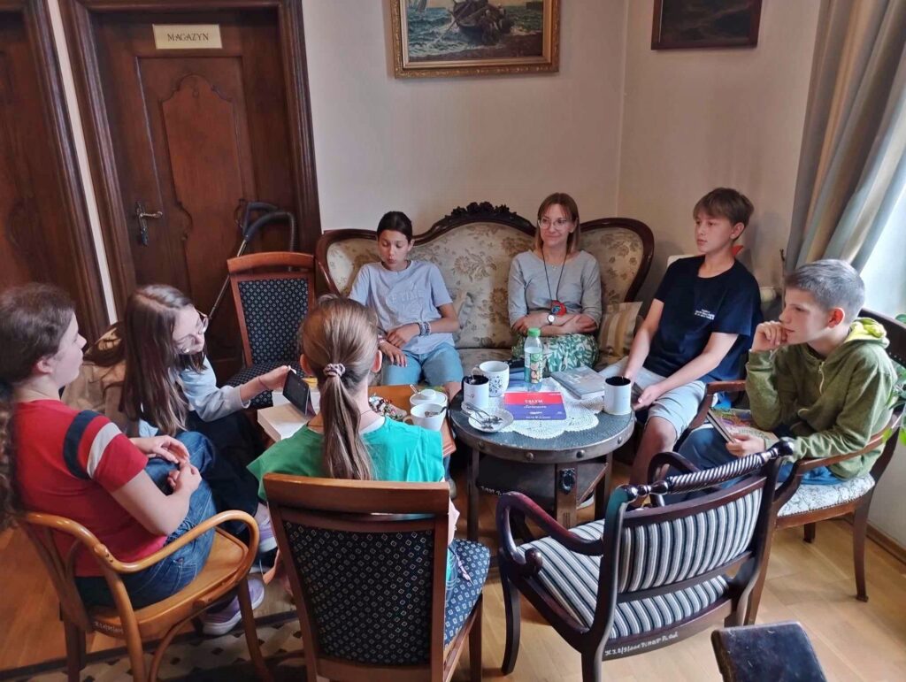 Przy 2 stolikach siedzą starsze dzieci i bibliotekarka. Na stolikach leżą książki, stoją kubki oraz słodycze.