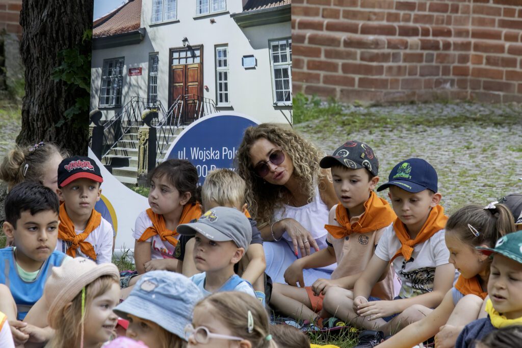 Na rozpostartych na trawie kocach siedzą dzieci. Między nimi nauczycielka. Za nimi baner przedstawiający budynek biblioteki.