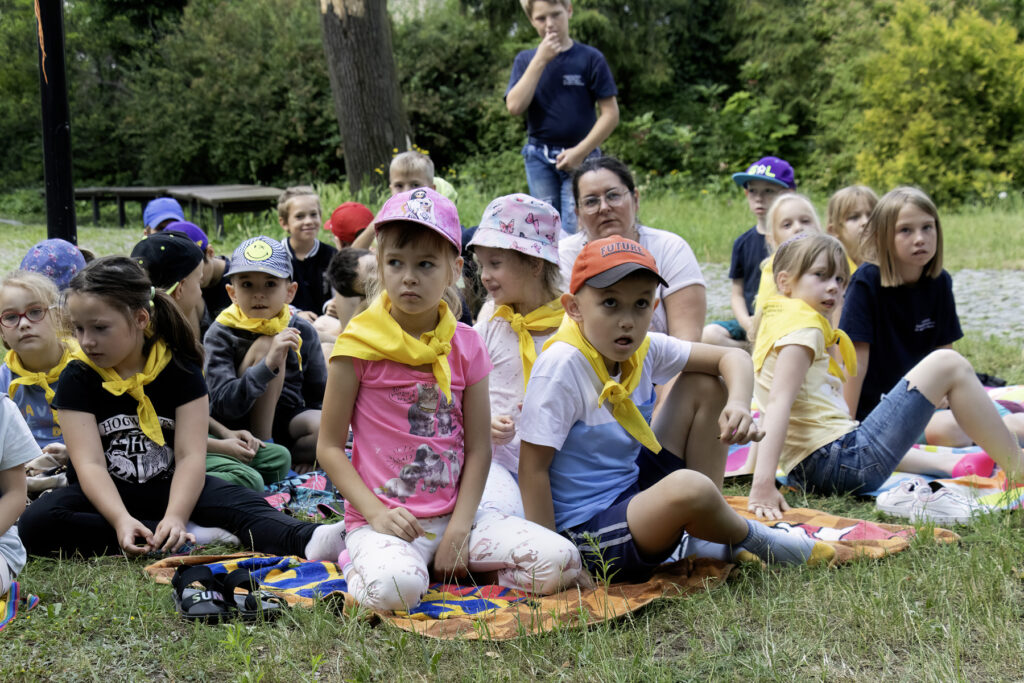 Na rozpostartych na trawie kocach siedzi duża grupa dzieci. 