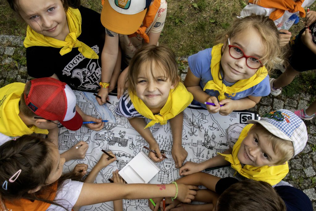 Dzieci malują mazakami zadrukowaną biała matę. Na macie rysunki: drzew,domów, wieżowców. kilkoro dzieci patrzy w  stronę osoby fotografującej i uśmiecha się.