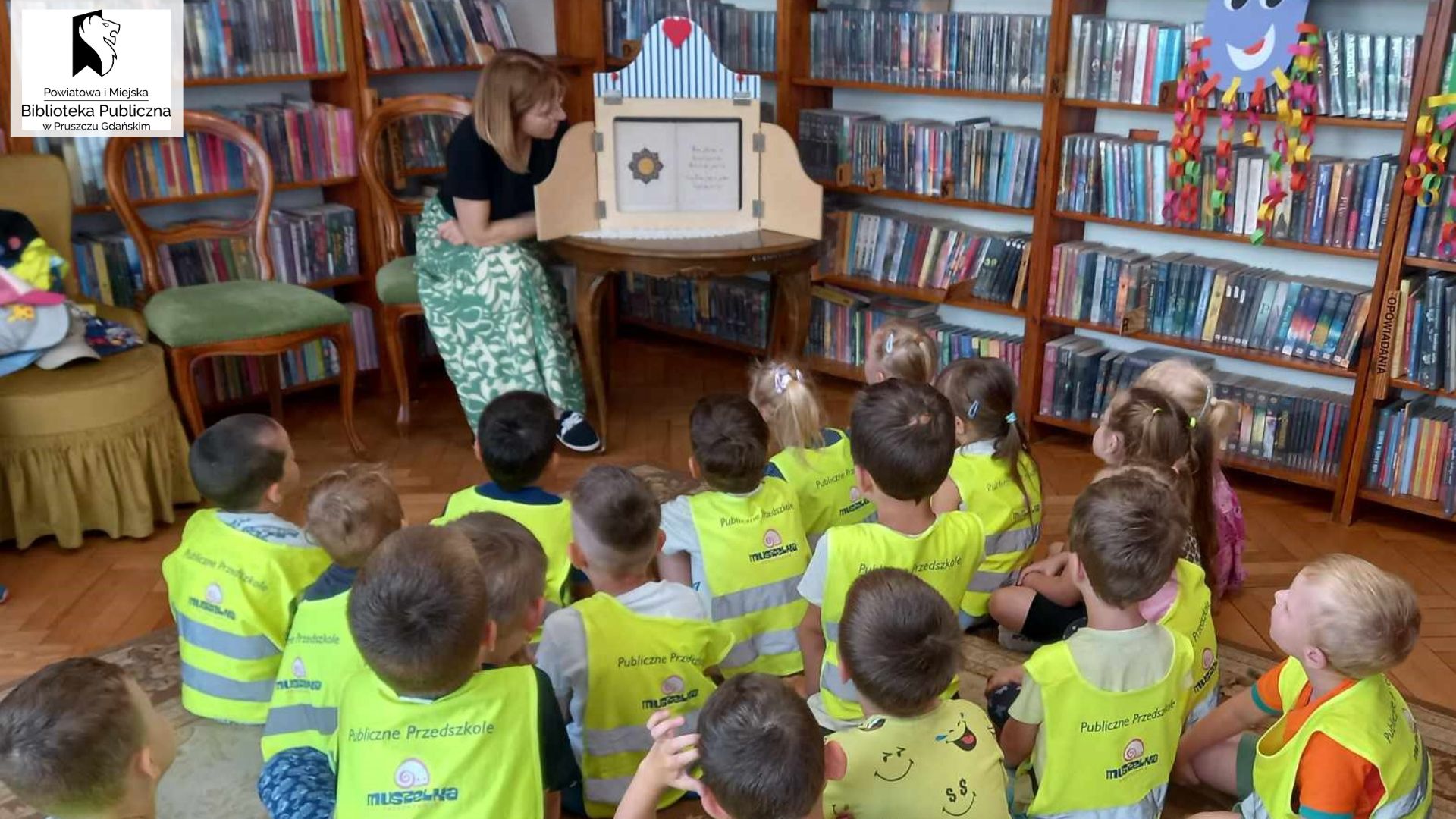 Bibliotekarka siedzi przy stoliku, przy otwartej skrzynce do teatrzyku kamishibai. Naprzeciwko niej siedzą dzieci na dywanie .