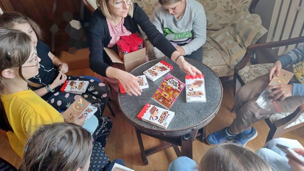 Wkoło stolika siedzą dzieci i bibliotekarka. Na stoliku bibliotekarka kładzie czekoladki. Na środku leży książka.