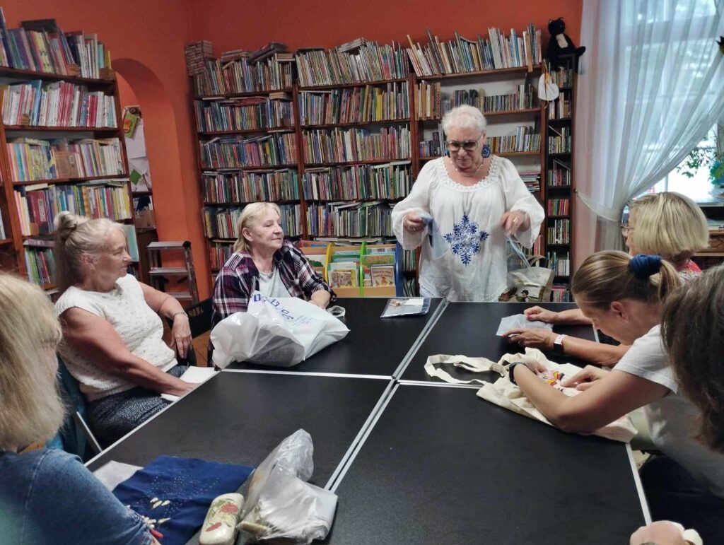 W sali bibliotecznej, przy stołach siedzi 6 kobiet. Wszystkie patrzą w stronę Anny Prill, która coś mówi. 