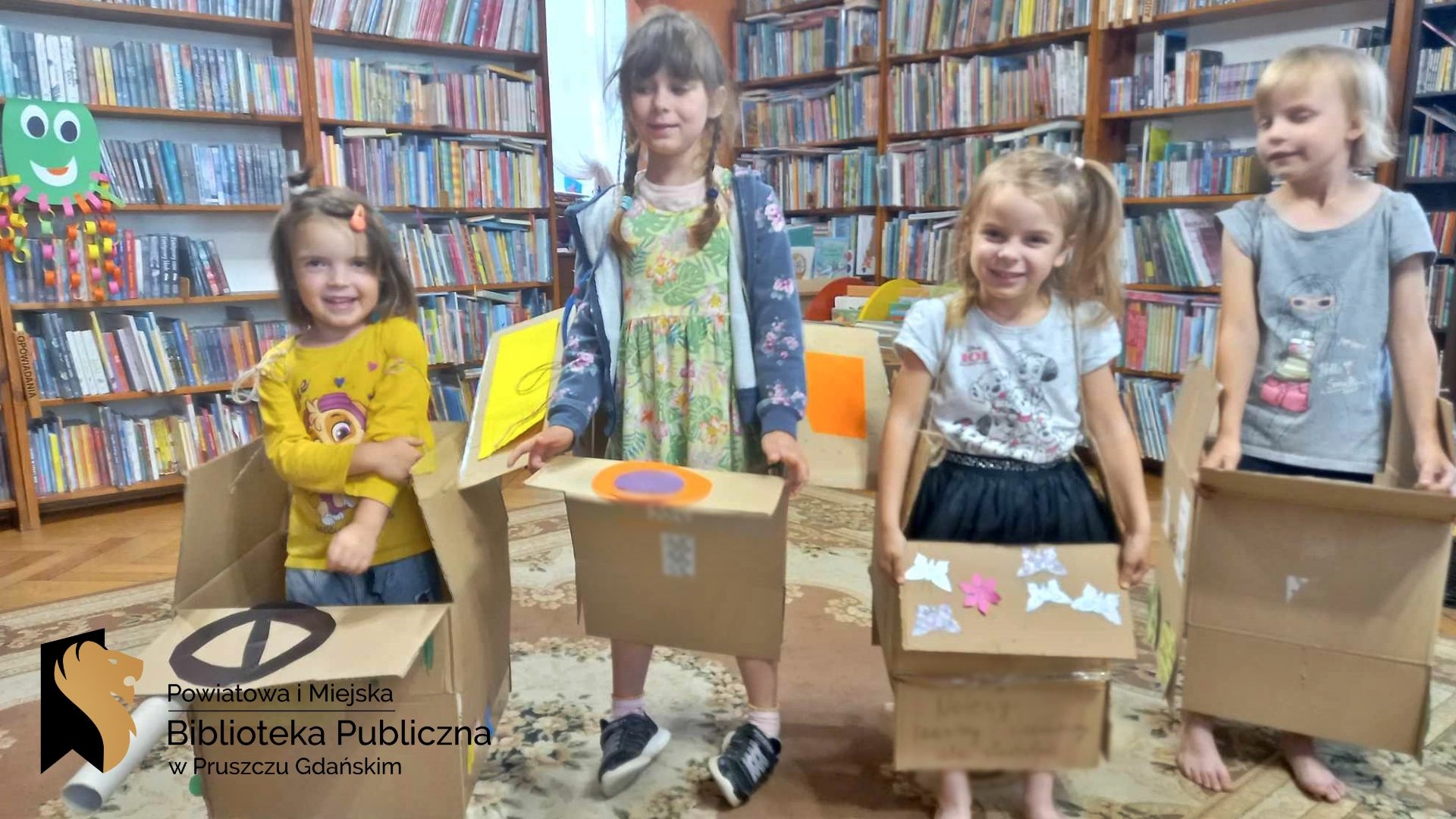 W sali bibliotecznej stoją 4 dziewczynki. dziewczynki stoją w kartonach ozdobionych mazakami i naklejkami.