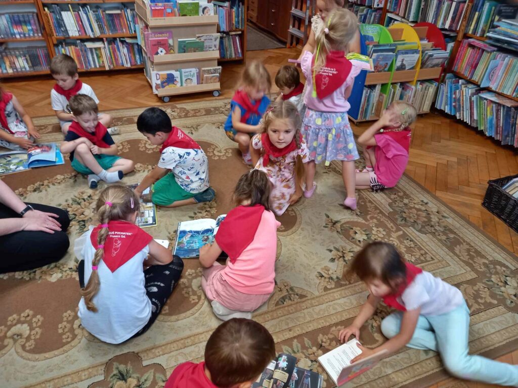 Na dywanie, w sali bibliotecznej siedzą dzieci i oglądają książki.