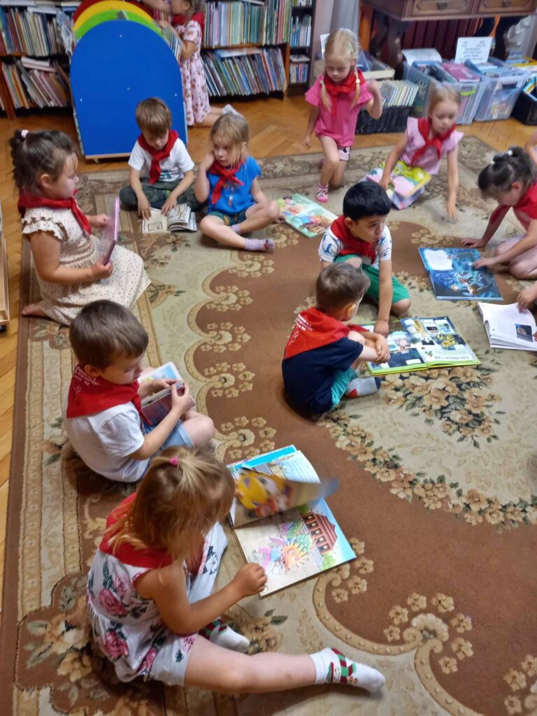 Na dywanie, w sali bibliotecznej siedzą dzieci i oglądają książki.