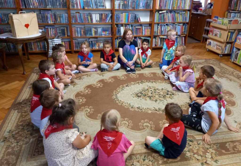 W sali bibliotecznej, na dywanie, siedzą dzieci. Dzieci patrzą na bibliotekarkę, która coś mówi. Na nimi, na tle regałów, stoi drewniany teatrzyk kamishibai.