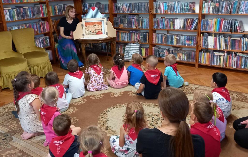 Na dywanie w okręgu siedzą dzieci. Dzieci patrzą na bibliotekarkę, która wyciąga 1 ilustrację i pokazuje 2 ilustrację, która umieszczona jest w teatrzyku kamishibi.  W tle regały z książkami.