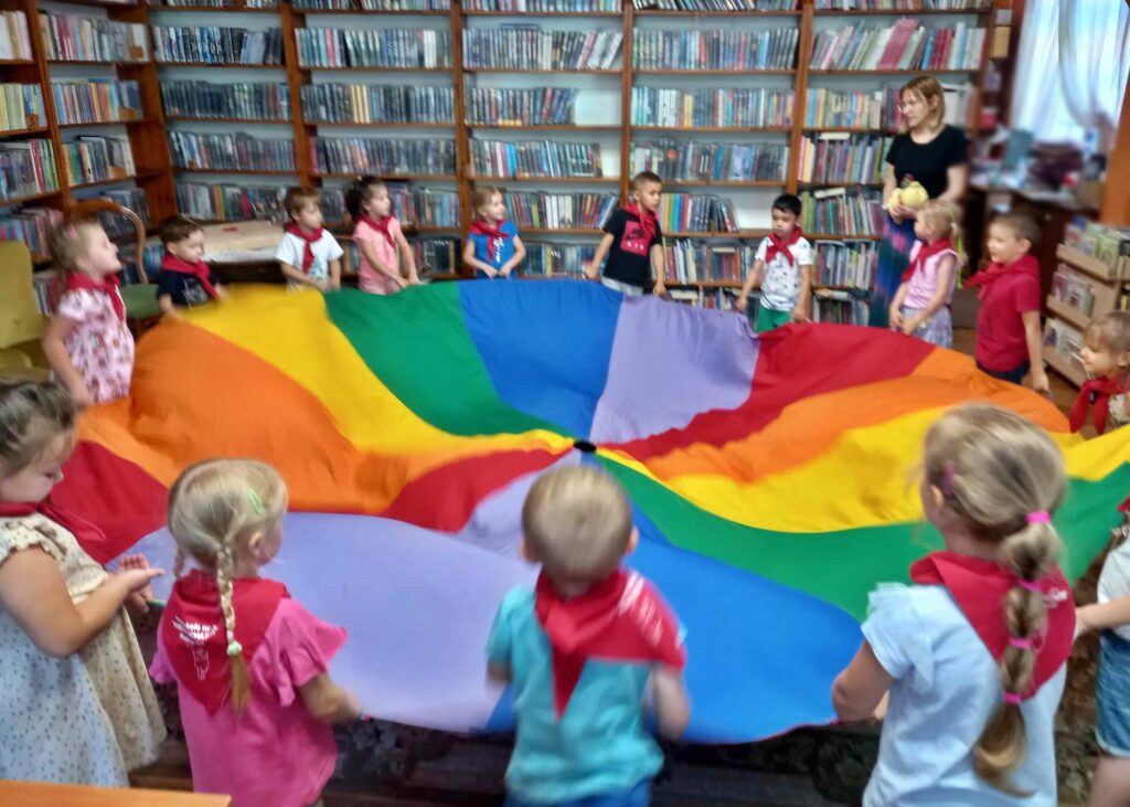 Dzieci stoją w okręgu i trzymają w rękach dużą, kolorową chustę Klanzy. Obok dzieci stoi bibliotekarka, która trzyma w rękach żółtego misia. W tle regały z książkami.