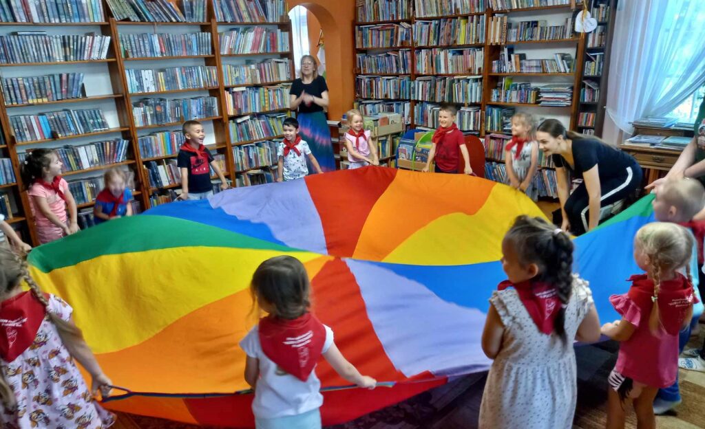 Dzieci stoją w okręgu i poruszają dużą, kolorową chustę Klanzy. Obok dzieci stoi bibliotekarka i coś mówi. W tle regały z książkami.