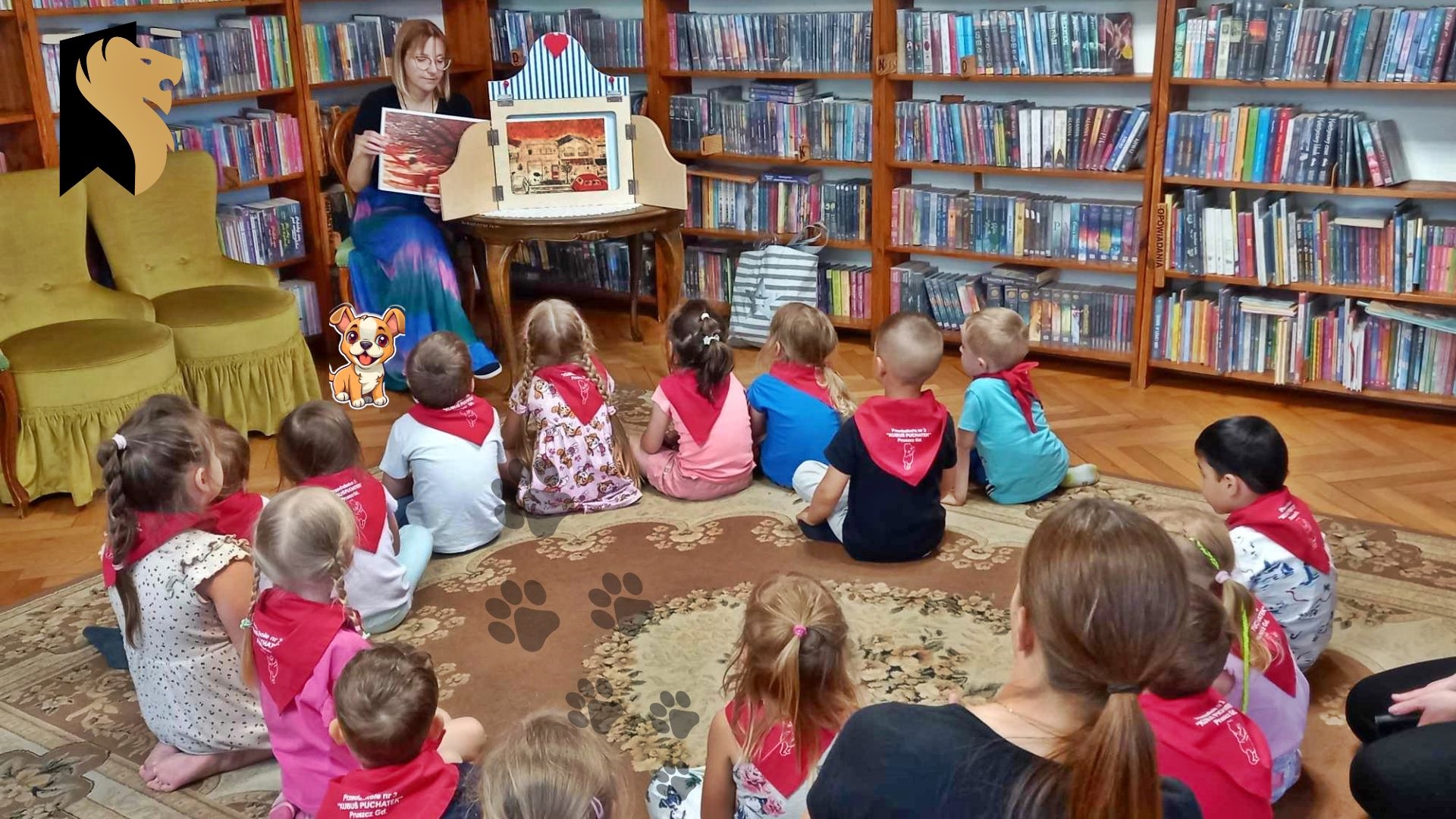 Na dywanie w okręgu siedzą dzieci. Dzieci patrzą na bibliotekarkę, która wyciąga 1 ilustrację i pokazuje 2 ilustrację, która umieszczona jest w teatrzyku kamishibi. W tle regały z książkami.