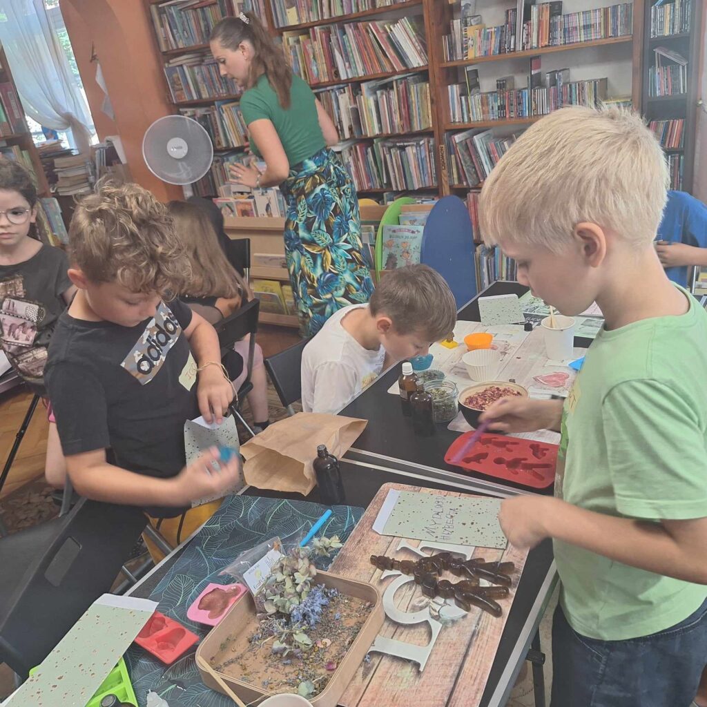 Sala biblioteczna. Wzdłuż sali rozstawione w 2 rzędach stoliki. Na stolikach olejki zapachowe, suszone kwiaty, różne foremki. Przy stolikach stoją i siedzą dzieci. 