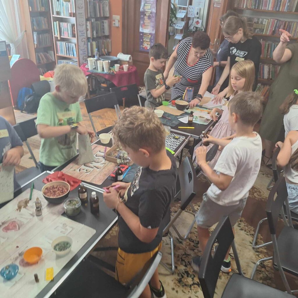 Sala biblioteczna. Wzdłuż sali rozstawione w 2 rzędach stoliki. Na stolikach olejki zapachowe, suszone kwiaty, różne foremki. Przy stolikach stoją i siedzą dzieci. 