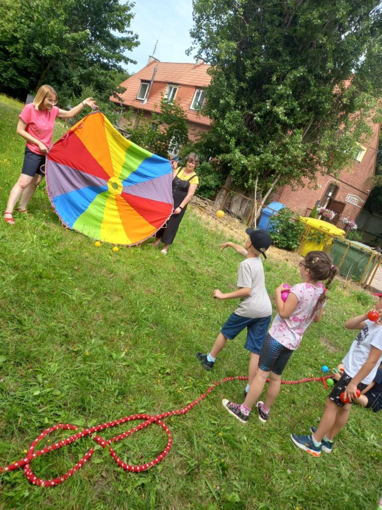 2 bibliotekarki trzymają rozłożoną pionowo chustę Klanzy. Przed nimi stoi 3 dzieci z piłeczkami w ręce. 