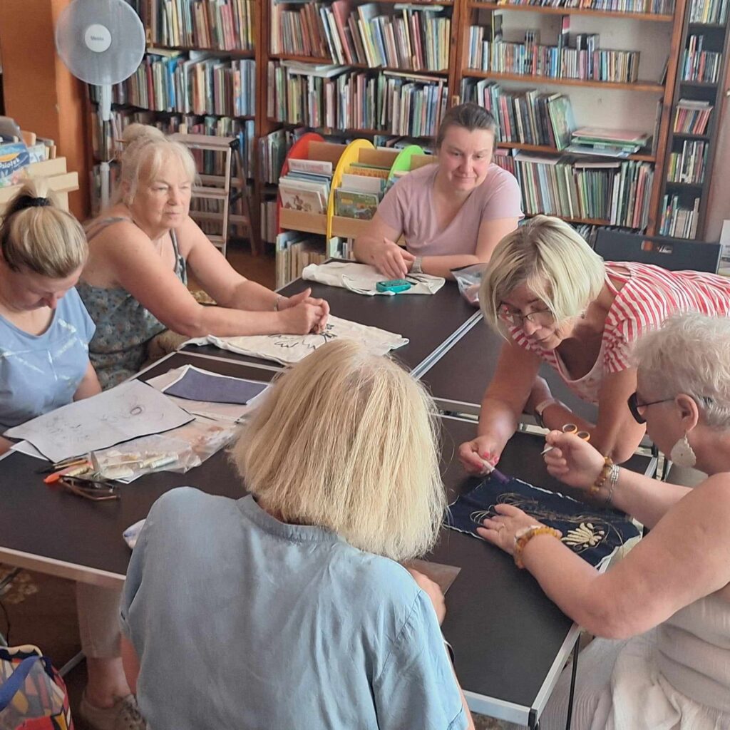 Sala biblioteczna. Przy stolikach siedzi 5 kobiet. 6 kobieta pochyla się na inną. Przed kobietami leżą materiały oraz kartki ze wzorami do  haftowania.