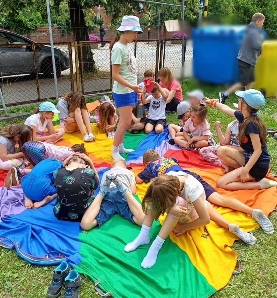 Kolorowa chusta Klanzy leży na trawie. Na chuście siedzą dzieci i mają głowy spuszczone w dół. Na środku chusty stoi 1 dziecko i patrzy w stronę drugiego dziecka, które wskazuje palcem na dziecko ze spuszczoną głową. 
