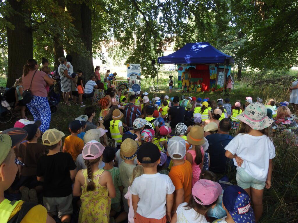 Na polanie, na kocach, siedzi duża grupa dzieci. Dzieci patrzą w stronę  stojącego na scenie aktora.