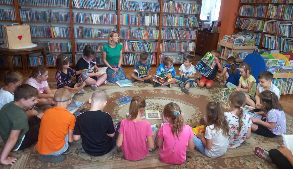 Sala biblioteczna. Na dywanie w okręgu siedzą dzieci i bibliotekarka. Bibliotekarka coś mówi. Dzieci trzymają w rękach książki i czasopisma.