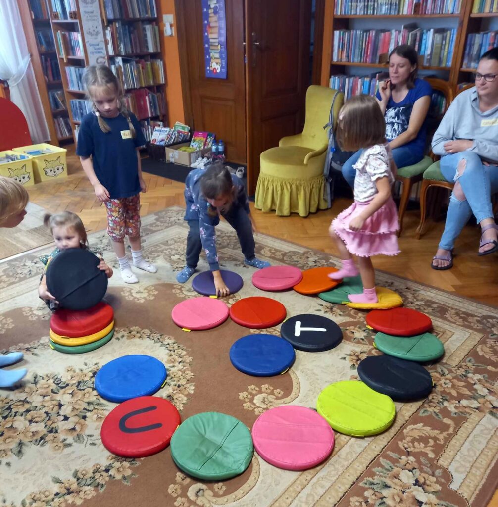 Na dywanie leżą kolorowe maty do siedzenia. Na kilku matach są literki. Po matach chodzą dzieci.